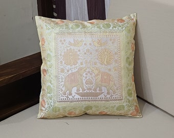 May include: A square decorative pillow with a detailed pattern. The pillow features a central design of two elephants facing each other, with peacocks and birds above. The color palette includes cream, gold, and peach tones, with floral accents around the border.