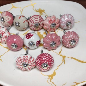 K&ouml;nnte beinhalten: Eine Sammlung von zw&ouml;lf runden und blumenf&ouml;rmigen Schrankkn&ouml;pfen mit verschiedenen rosa, wei&szlig;en und roten Mustern. Jeder Knopf besteht aus Keramik mit einer zentralen silberfarbenen Metallschraube und einer kleinen runden Basisplatte.