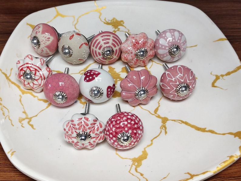 K&ouml;nnte beinhalten: Eine Sammlung von zw&ouml;lf runden und blumenf&ouml;rmigen Schrankkn&ouml;pfen mit verschiedenen rosa, wei&szlig;en und roten Mustern. Jeder Knopf besteht aus Keramik mit einer zentralen silberfarbenen Metallschraube und einer kleinen runden Basisplatte.
