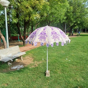 May include: A white and purple patterned umbrella with a fringe border. The umbrella is open and standing on a grassy lawn in a park.