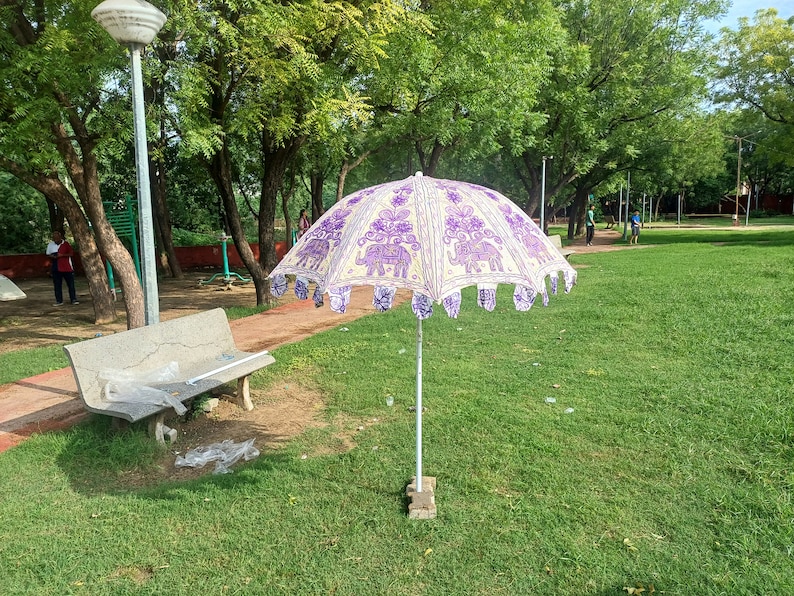 May include: A white and purple patterned umbrella with a fringe border. The umbrella is open and standing on a grassy lawn in a park.