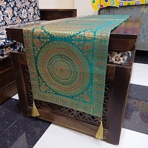 Op de afbeelding: Een teal en goudkleurige tafelloper met een centraal cirkelvormig ontwerp en decoratieve kwastjes. De loper is gedrapeerd over een donkere houten tafel, wat een vleugje elegantie aan de kamer toevoegt. De tafel staat voor een bank met patronen.