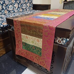 May include: A pink, gold, and green table runner with a floral pattern. The runner is on a dark wood table with carved details.