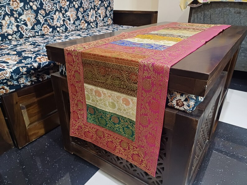 May include: A pink, gold, and green table runner with a floral pattern. The runner is on a dark wood table with carved details.