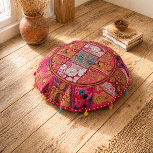 Peut inclure: Un coussin de sol rond rose avec des motifs de patchwork complexes et des broderies dorées. Le coussin présente une variété de couleurs, notamment rose, orange, bleu et blanc, et est orné de petits pompons colorés sur le bord. Le coussin est posé sur un sol en bois.