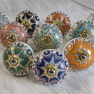 Op de afbeelding: Tien keramische knoppen met ingewikkelde bloemmotieven in verschillende kleuren, waaronder turquoise, groen, oranje, roze en blauw. Elke knop heeft een gouden midden met een reliëfdesign.