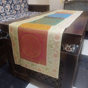 May include: A multi-colored table runner with a cream border and intricate floral designs. The runner is made of a soft, luxurious fabric and features a square, red and gold patterned center.