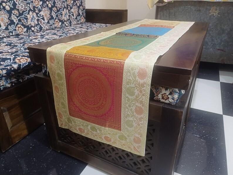 May include: A multi-colored table runner with a cream border and intricate floral designs. The runner is made of a soft, luxurious fabric and features a square, red and gold patterned center.