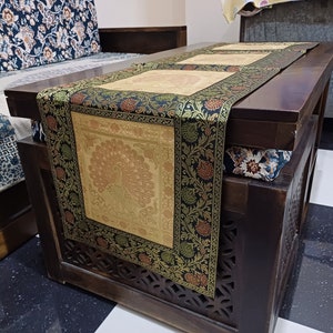 May include: A black and gold table runner with a peacock design. The runner is made of brocade fabric and has a fringed edge. The runner is on a wooden table with a carved design.