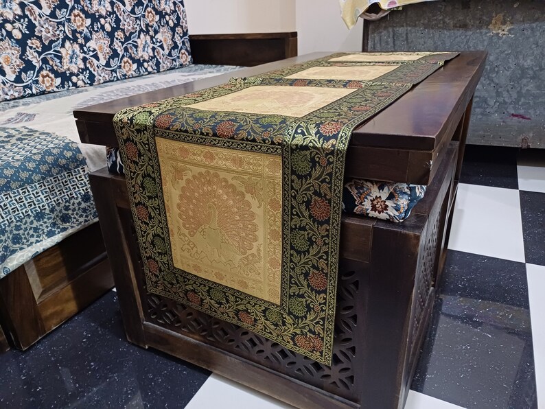 May include: A black and gold table runner with a peacock design. The runner is made of brocade fabric and has a fringed edge. The runner is on a wooden table with a carved design.