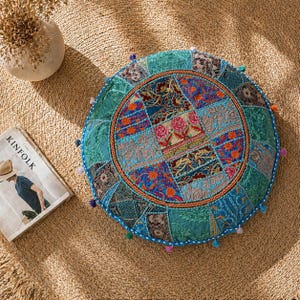 May include: Round, patchwork floor cushion in shades of teal, blue, and orange. The cushion features a central circular design with floral and geometric patterns. The edges are trimmed with small, colorful pom-poms. A magazine with the word "KINFOLK" is visible.