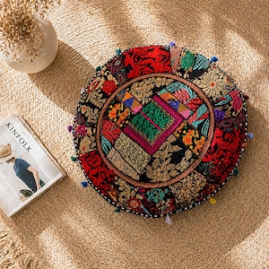 May include: A round, colorful floor cushion with a patchwork design. The cushion features a black background with red, gold, green, and blue embroidered details. Small tassels are attached around the edge. A magazine and a vase are in the background.