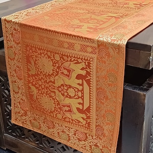Puede incluir: Un camino de mesa naranja con un diseño bordado en oro de elefantes y pavos reales. El camino de mesa está sobre una mesa de madera con un diseño tallado.