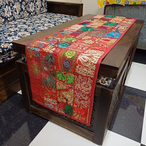 K&ouml;nnte beinhalten: Ein roter und goldfarbener bestickter Tischl&auml;ufer mit Patchwork-Muster. Der L&auml;ufer liegt auf einem dunklen Holztisch mit einem floralen Kissen.