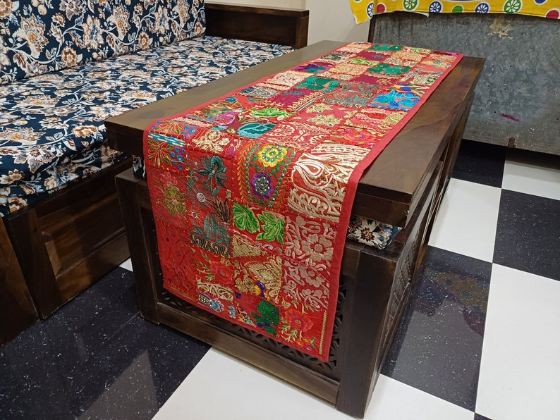 K&ouml;nnte beinhalten: Ein roter und goldfarbener bestickter Tischl&auml;ufer mit Patchwork-Muster. Der L&auml;ufer liegt auf einem dunklen Holztisch mit einem floralen Kissen.