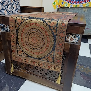 Peut inclure: Chemin de table décoratif avec un fond noir et un motif circulaire complexe doré et rouge. Le chemin de table a des glands dorés à chaque extrémité et est drapé sur une table en bois. La table a un motif sculpté décoratif.