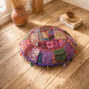 May include: A round floor cushion with a patchwork design in shades of purple, pink, and gold. The cushion has a purple border and is adorned with small tassels in various colors. The cushion is placed on a wooden floor.