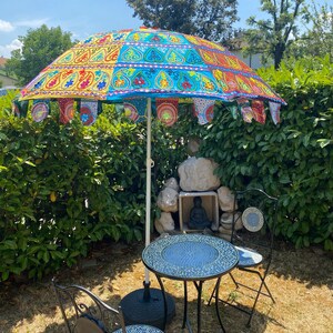 May include: A patio set consisting of a round mosaic tile table and two matching chairs, positioned under a large, colorful parasol. The parasol features vibrant panels of yellow, blue, and red fabric with intricate embroidery and hanging fabric flags.