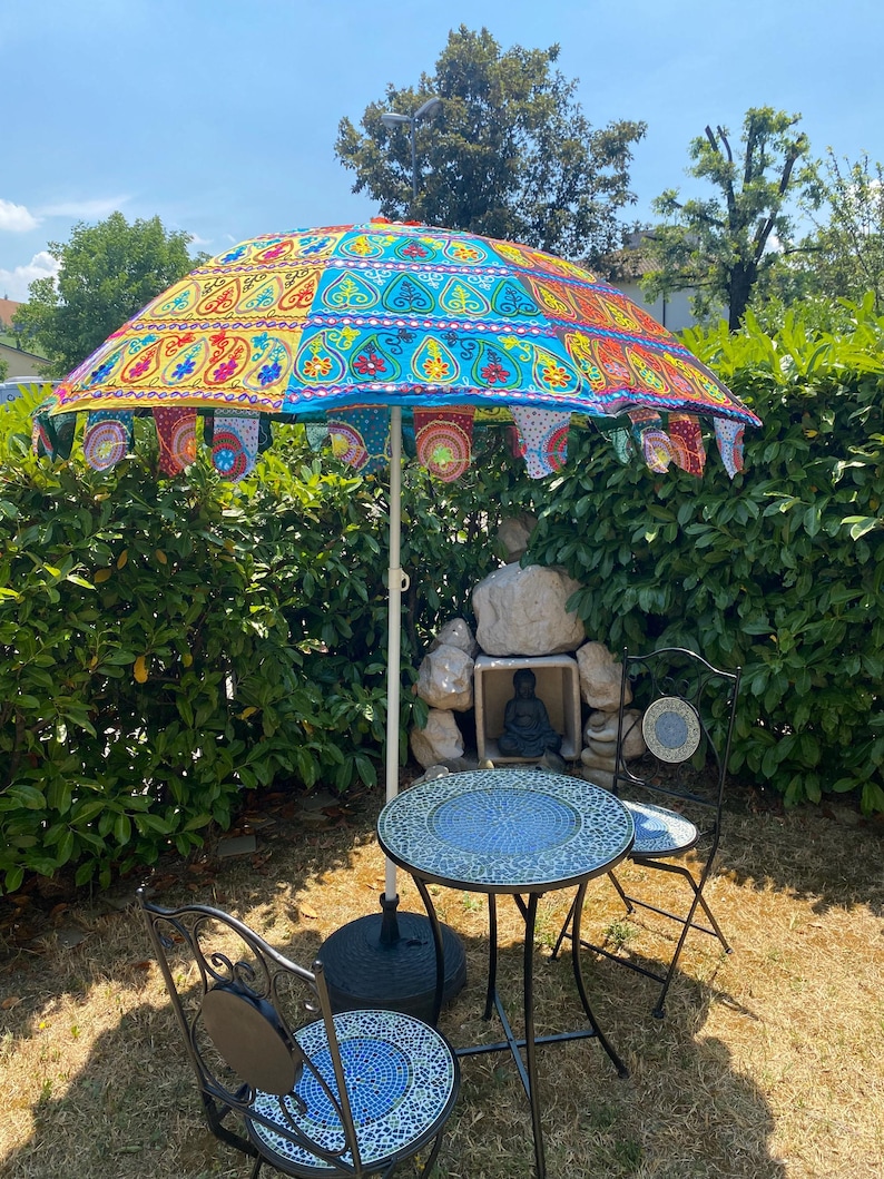 May include: A patio set consisting of a round mosaic tile table and two matching chairs, positioned under a large, colorful parasol. The parasol features vibrant panels of yellow, blue, and red fabric with intricate embroidery and hanging fabric flags.