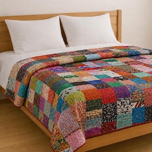 Op de afbeelding: Een patchwork quilt met een levendige mix van kleuren en patronen, waaronder rode, oranje, groene en blauwe vierkanten. De quilt ligt op een bed met witte kussens en een houten hoofdbord, wat een gezellige sfeer creëert.