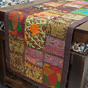 K&ouml;nnte beinhalten: Ein brauner Tischl&auml;ufer mit Patchwork-Design, der bestickte Blumen- und Paisley-Muster in den Farben Lila, Orange, Gr&uuml;n und Braun zeigt. Der L&auml;ufer hat einen dunkelbraunen Rand.