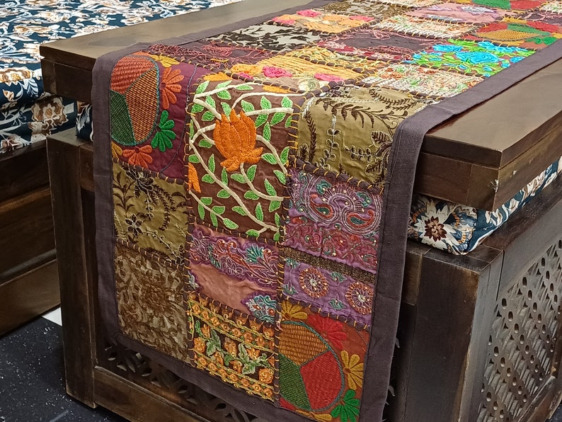 K&ouml;nnte beinhalten: Ein brauner Tischl&auml;ufer mit Patchwork-Design, der bestickte Blumen- und Paisley-Muster in den Farben Lila, Orange, Gr&uuml;n und Braun zeigt. Der L&auml;ufer hat einen dunkelbraunen Rand.