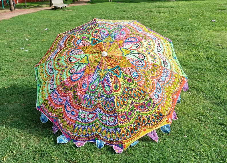 May include: A circular parasol featuring a vibrant, multi-colored mandala pattern with intricate paisley and floral motifs in shades of yellow, pink, blue, green, and orange. The edge is finished with small, decorative fabric triangular accents.