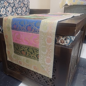 Può includere: Un runner da tavolo multicolore con motivi floreali in tonalità di blu, rosa, verde e oro. Il runner è posizionato su un tavolo in legno scuro con intagli intricati.