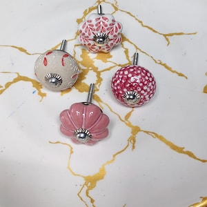 K&ouml;nnte beinhalten: Vier dekorative Schrankkn&ouml;pfe mit Metallbeschl&auml;gen. Einer ist eine wei&szlig;e Blumenform mit roten Mustern, einer ist ein runder beiger Knopf mit roten Blumendetails, einer ist ein runder rosa Knopf mit wei&szlig;en Punkten und einer ist ein rosa Blumenknopf.