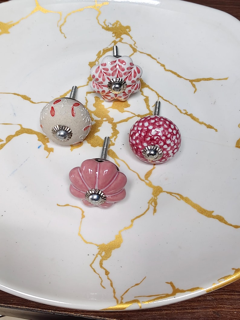 K&ouml;nnte beinhalten: Vier dekorative Schrankkn&ouml;pfe mit Metallbeschl&auml;gen. Einer ist eine wei&szlig;e Blumenform mit roten Mustern, einer ist ein runder beiger Knopf mit roten Blumendetails, einer ist ein runder rosa Knopf mit wei&szlig;en Punkten und einer ist ein rosa Blumenknopf.