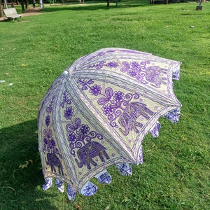May include: A white and purple umbrella with an embroidered design of elephants and flowers. The umbrella is open and lying on a grassy lawn.