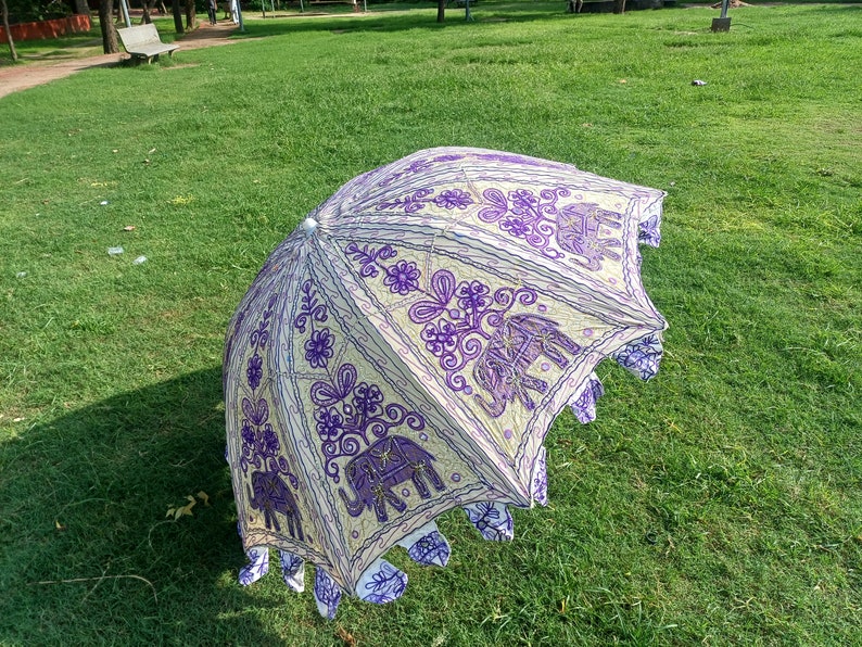 May include: A white and purple umbrella with an embroidered design of elephants and flowers. The umbrella is open and lying on a grassy lawn.