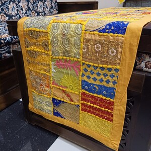 K&ouml;nnte beinhalten: Ein gelber Tischl&auml;ufer mit Patchwork-Design in verschiedenen Farben und Texturen. Der L&auml;ufer ist aus Stoff mit Stickereien und mit verschiedenen Mustern verziert, darunter florale Motive, geometrische Formen und Paisley-Muster.