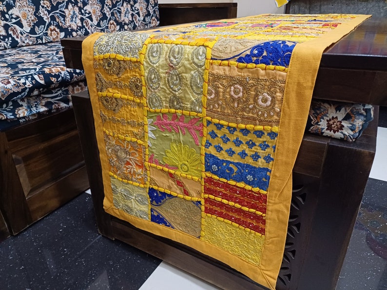 K&ouml;nnte beinhalten: Ein gelber Tischl&auml;ufer mit Patchwork-Design in verschiedenen Farben und Texturen. Der L&auml;ufer ist aus Stoff mit Stickereien und mit verschiedenen Mustern verziert, darunter florale Motive, geometrische Formen und Paisley-Muster.