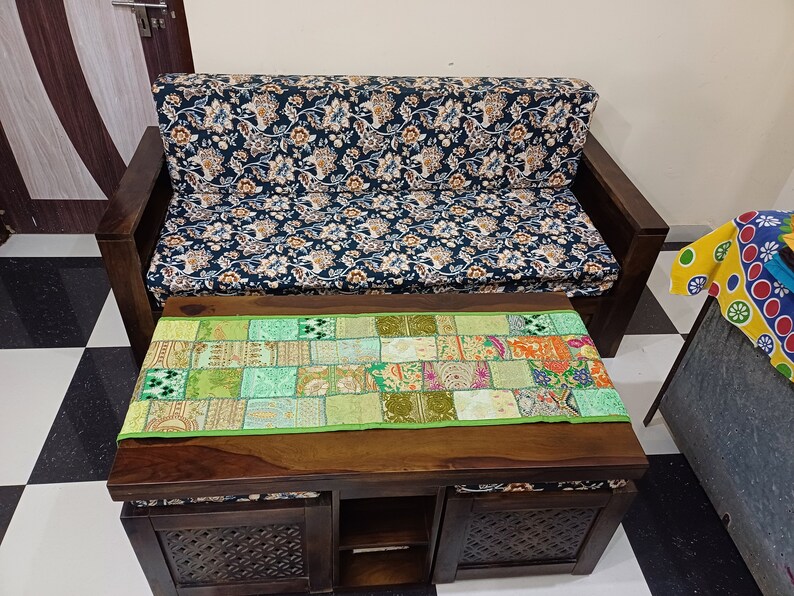 K&ouml;nnte beinhalten: Ein dunkles Holzsofa mit einem floral gemusterten Stoffbezug und ein passender dunkler Holz-Couchtisch mit einem gr&uuml;nen Patchwork-L&auml;ufer. Der Couchtisch hat zwei Schubladen und ein offenes Regal.
