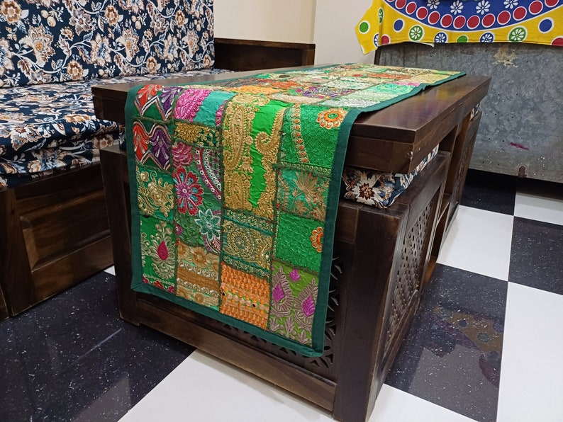 K&ouml;nnte beinhalten: Ein gr&uuml;ner Patchwork-Tischl&auml;ufer mit bunten Stickereien und goldenen Akzenten. Der L&auml;ufer liegt auf einem dunklen Holztisch mit einem geschnitzten Design.