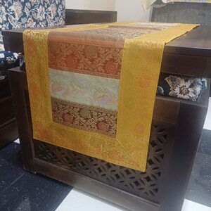 May include: A multi-colored table runner with a gold border, featuring a floral pattern. The runner is placed on a dark wood table with intricate carvings.