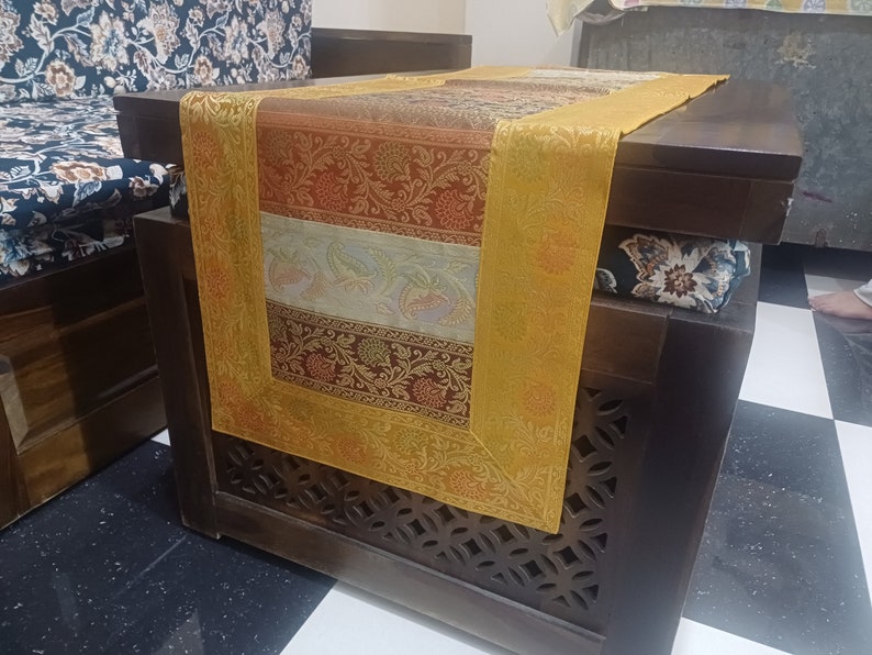 May include: A multi-colored table runner with a gold border, featuring a floral pattern. The runner is placed on a dark wood table with intricate carvings.