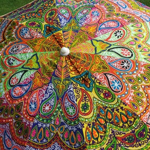 May include: A parasol featuring a vibrant, multi-colored fabric canopy with intricate embroidery and mirror work. The design displays a radial pattern of paisley motifs, floral shapes, and geometric lines in shades of orange, yellow, green, blue, and pink, centered around a white circular finial.