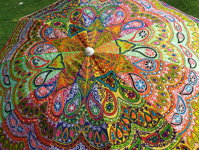 May include: A parasol featuring a vibrant, multi-colored fabric canopy with intricate embroidery and mirror work. The design displays a radial pattern of paisley motifs, floral shapes, and geometric lines in shades of orange, yellow, green, blue, and pink, centered around a white circular finial.