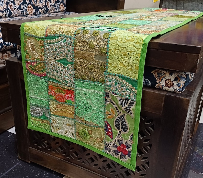 K&ouml;nnte beinhalten: Ein gr&uuml;ner Patchwork-Tischl&auml;ufer mit Stickereien und Perlenbesatz. Der L&auml;ufer besteht aus verschiedenen Stoffen und hat einen Fransenrand.