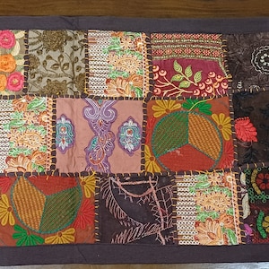 May include: A colourful patchwork table runner with embroidered floral and geometric designs in shades of brown, red, green, blue, and orange.