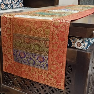 May include: A red, gold, and green patterned table runner with a floral design. The runner is on a dark wood table with a carved design.