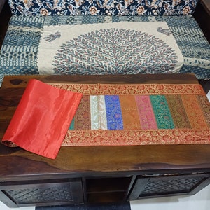 以下が含まれることがあります： カラフルなテーブルランナーと丸められた赤いシルクのプレースマットが置かれた木製のコーヒーテーブル。 ランナーは、さまざまな色と複雑なデザインの垂直ストライプが特徴です。 パターンのクッションとソファが背景にあります。