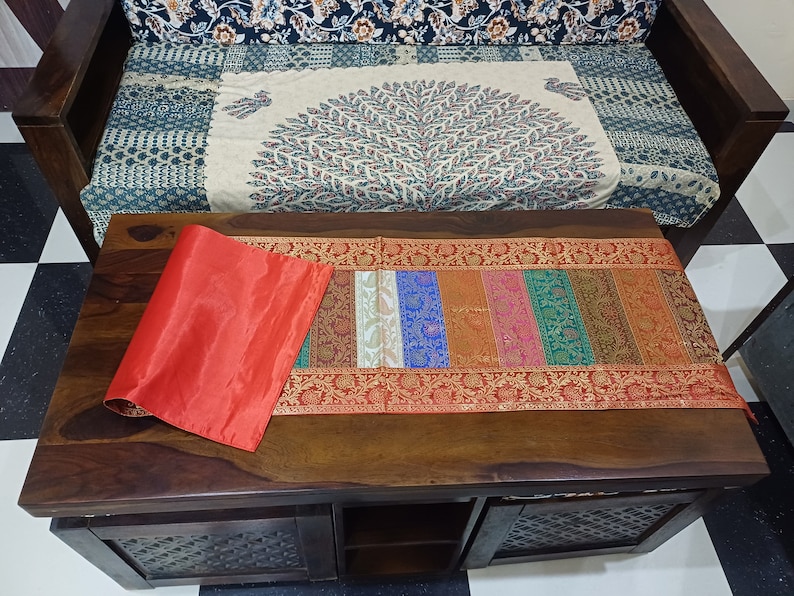 以下が含まれることがあります： カラフルなテーブルランナーと丸められた赤いシルクのプレースマットが置かれた木製のコーヒーテーブル。 ランナーは、さまざまな色と複雑なデザインの垂直ストライプが特徴です。 パターンのクッションとソファが背景にあります。