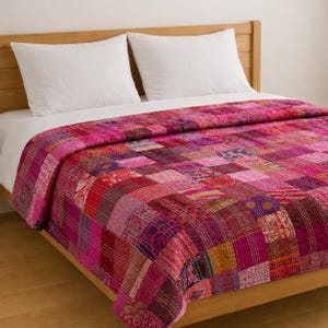 May include: A patchwork quilt in shades of pink, red, and purple covers a bed. The quilt is made of various fabric squares stitched together. White pillows rest on the bed. The quilt's design is a mix of patterns and textures.
