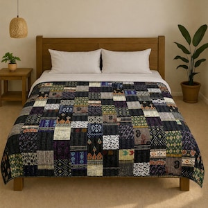 Op de afbeelding: Een patchwork quilt met verschillende patronen in zwart, paars, groen en beige bedekt een bed. De quilt ligt op een houten bedframe met witte kussens en een wit laken. Een kleine bijzettafel en een plant in een pot zijn ook zichtbaar.