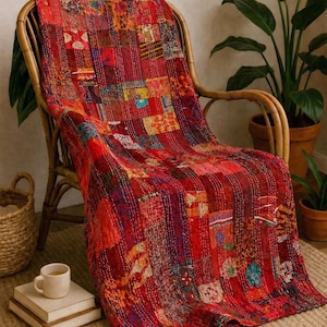 Op de afbeelding: Een levendige rode patchworkquilt over een rotan stoel. De quilt heeft een verscheidenheid aan kleurrijke stoffen vierkanten die met witte draad aan elkaar zijn gestikt. De stoel staat in een gezellige kamer met planten en een geweven vloerkleed.
