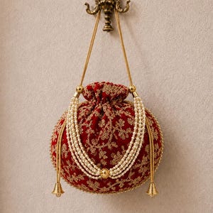 Può includere: Una borsa a sacchetto in velluto rosso scuro con chiusura a coulisse e ricami floreali dorati. La borsa è decorata con diversi fili di perle bianche finte drappeggiate sul davanti e nappe in metallo dorato alla base dei cordini.