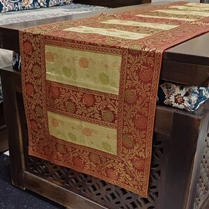 May include: A red and gold table runner with a floral pattern. The runner is made of a brocade fabric and has a fringed edge. The runner is on a wooden table with a carved design.